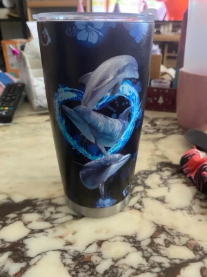 Svart termosmugg med delfinmotiv - Cool svart termosmugg i rostfritt stål med färgglatt tryck av tre delfiner och ett blått hjärta. Muggen har genomskinligt lock och blommönster i bakgrunden. Perfekt för att ta med drycken på språng.