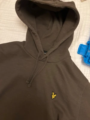 Brun hoodie från Lyle & Scott - Säljer en brun hoodie från Lyle & Scott med klassisk gul logga på bröstet. Hoodien har dragsko i huvan och är tillverkad i mjukt bomullsmaterial. Perfekt för dig som gillar enkel och stilren streetwear. Hör av er vid mer info storlek xs!