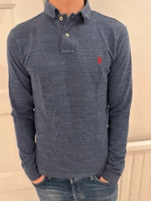 Blå långärmad henley från Ralph Lauren - Mörkblå långärmad henley från Ralph Lauren med klassisk krage och två knappar vid halsen. Skjortan har en liten röd broderad logga på bröstet och är gjord i mjuk bomull. Perfekt till jeans för en avslappnad men snygg look.