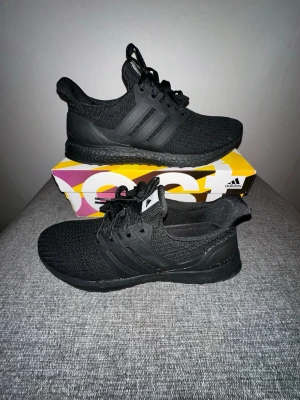 Svarta Adidas Ultra Boost sneakers - Säljer ett par helsvarta Adidas Ultra Boost sneakers med stickad ovandel och klassiska tre ränder på sidan. Skorna har svart sula och snörning, samt Adidas-logga på plösen. Perfekta för dig som gillar stilrena och bekväma sneakers.
