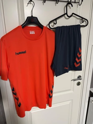 Röd träningsset från Hummel - Säljer ett sportigt träningsset från Hummel med röd t-shirt och mörkblå shorts. T-shirten har svarta Hummel-loggor på bröstet och sidorna, och shortsen har matchande detaljer. Perfekt för fotboll eller gym. Materialet är lätt och andas.