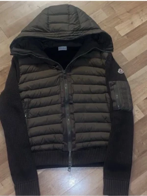 Oliv grön Moncler pufferjacka med huva - Stilren grön pufferjacka från Moncler med stickade ärmar och vadderad kropp. Jackan har huva, dragkedja framtill och en ficka med dragkedja på ärmen. Perfekt för kyliga dagar och snygg till vardags. QR kod och NFC.