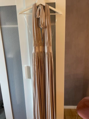 Champagnefärgad långklänning med knyt - Elegant långklänning i skimrande champagnefärg med djup v-ringning och bred midjedetalj. Klänningen har knyt i nacken och ett mjukt, fallande tyg som ger en lyxig känsla. Perfekt för speciella tillfällen. Har dock små gräns fläckar längst ner på klänningen men inget som syns alls💕Finns ingen storlek på klänning men passar mellan storlekarna Xs-S❤️