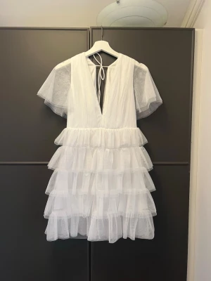 Vit volangklänning med v-ringning - Supersöt vit kortklänning med flera lager volanger och djup v-ringning. Klänningen har transparenta korta ärmar och knytning i nacken. Perfekt för dig som vill ha en luftig och romantisk look. Väldigt populär! Perfekt till student!