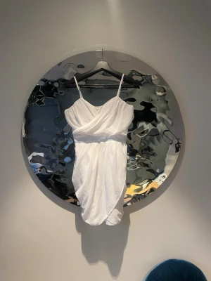 Vit omlottklänning med smala band - Säljer en vit kortklänning med omlottdesign och smala axelband. Klänningen har en draperad look och v-ringad urringning, vilket ger en elegant och modern känsla. Perfekt för sommarens alla tillfällen!