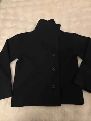 Mörkblå kappa - Mörkblå kappa med tre knappar framtill. Kappan har raka ärmar och en minimalistisk design som passar dig som gillar klassiska plagg. Materialet ser ut att vara kraftigt och håller formen snyggt. Perfekt för dig som vill ha en enkel och cool look.