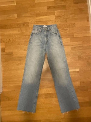 Ljusblåa raka jeans med rå kant - Säljer ett par ljusblå jeans med hög midja och raka ben. Jeansen har fem fickor och råa, fransiga benslut för en avslappnad look. Tillverkade i klassisk denim med en tvättad finish. Perfekta för dig som gillar en trendig och bekväm stil.