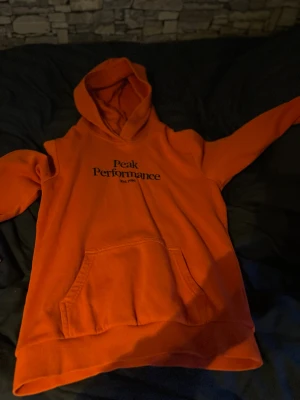 Orange hoodie från Peak Performance - Cool orange hoodie från Peak Performance med svart logga på bröstet. Tröjan har huva och en stor magficka framtill. Materialet är mjukt och fleeceliknande på insidan, perfekt för att hålla sig varm. Passar dig som gillar sportig och avslappnad stil.