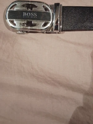 Svart skärp från BOSS med silverdetaljer - Snyggt svart skärp från BOSS med en bred, stilren rem i skinn och en stor, blank silverfärgad metallspänne med BOSS-loggan. Perfekt accessoar för att lyfta din outfit med en exklusiv touch.