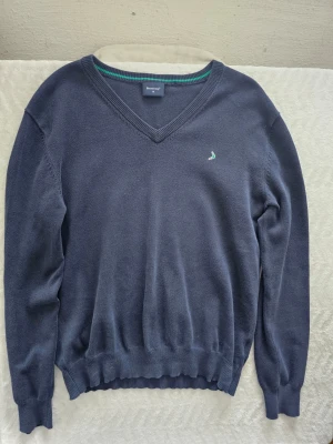 Mörkblå V-ringad tröja Boomerang - Klassisk mörkblå stickad tröja från Boomerang med V-ringning och diskret logga broderad på bröstet. Tröjan har ribbade muddar vid ärmslut och nederkant samt en grön rand på insidan av kragen. Perfekt till jeans eller chinos.