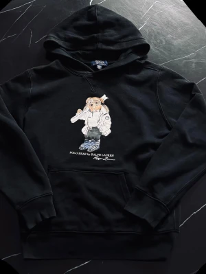 Ralph Lauren Polo Bear Hoodie Svart – Trendig & Populär - Säljer en snygg Polo Bear hoodie från Ralph Lauren. Klassisk svart hoodie med den ikoniska Polo Bear-designen på framsidan. Väldigt populär modell och passar perfekt till vardag eller streetwear. Storlek: 14–16 år (passar ungefär S) Färg: Svart Mått: • Längd: 58 cm • Bredd (armhåla till armhåla): 50 cm Skick: Mycket fint skick. Inga hål eller större fläckar vad jag kan se. Skickas eller möts upp om det passar. Skriv om du vill ha fler bilder eller mer info!