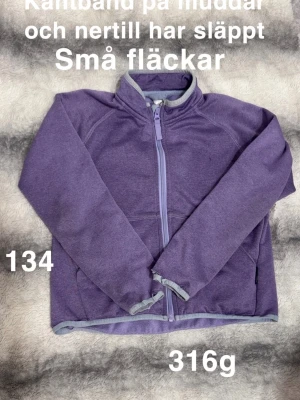 Lila tröja med dragkedja - Lila tröja med hel dragkedja och hög krage. Tröjan har långa ärmar och ljuslila kantband vid muddar och nederkant. 🌸 Kantbanden vid muddarna samt där nere har släppt på vissa ställen. Några små fläckar. Några noppor. Storlek 134.