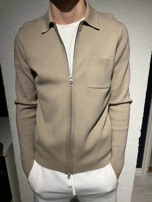 Beige ziptröja med krage - Snygg beige ziptröja med krage och bröstficka. Tröjan har en rak passform, långa ärmar och är tillverkad i ett mjukt, stickat material. Perfekt för en clean och stilren look.