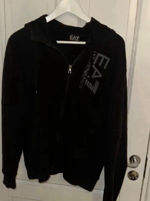Svart dress från EA7 Emporio Armani - Snygg svart hoodie och byxor från EA7 Emporio Armani med dragkedja framtill och stor logga på bröstet. Hoodien har huva och är tillverkad i mjukt material som känns skönt mot huden. Perfekt för en avslappnad och trendig look.