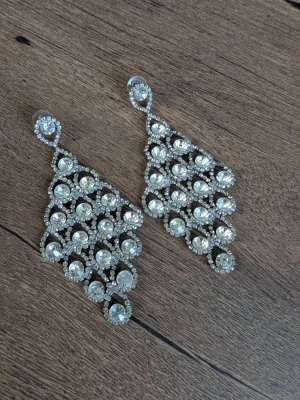 Silvriga statement örhängen med stenar - Säljer ett par stora, glittriga örhängen i silverfärg med massor av klara stenar i pärlform. Örhängena har en triangulär form och är täckta av gnistrande stenar som ger en riktigt lyxig känsla. Perfekta för dig som vill sticka ut och älskar bling!