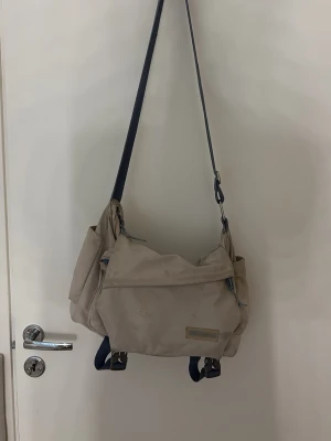 Beige axelväska från Xinding - Säljer en beige axelväska från Xinding med justerbar axelrem i mörkblått. Väskan har flera fack med dragkedja och detaljer i blått. Tillverkad i slitstark canvas, perfekt för dig som vill ha en praktisk och stilren väska till vardagen.