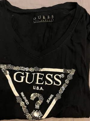 Svart långärmad topp från Guess - Snygg svart långärmad topp från Guess med v-ringning och ikoniskt triangellogotyp framtill dekorerad med pärlor och detaljer. Tillverkad i mjukt material som känns skönt mot huden. Perfekt för dig som vill ha en trendig och stilren look.