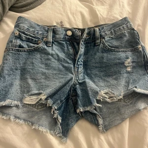 Blå denimshorts med slitningar - Säljer ett par blå denimshorts från 157 med råa kanter och slitna detaljer framtill. Klassisk femficksmodell med knappgylf och bälteshällor. Perfekta för sommaren och ger en avslappnad look.