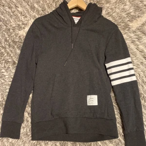 Mörkgrå hoodie från Thom Browne - Snygg mörkgrå hoodie från Thom Browne med klassiska vita ränder på ena ärmen och en liten patch framtill. Tröjan har huva med snörning och känguruficka. Perfekt för dig som gillar stilrena och sportiga plagg.