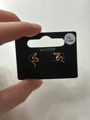 Guldiga ormörhängen från Montini - Säljer ett par snygga örhängen från Montini i form av små ormar. De är guldfärgade och har en glittrig yta som ger extra attityd till din look. Perfekta för dig som vill ha något unikt och trendigt i öronen.