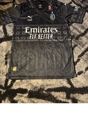 Svart AC Milan matchtröja Puma - Snygg svart AC Milan matchtröja från Puma med diskret mönster, vita loggor och Emirates Fly Better-tryck på bröstet. Klassisk sportig passform och officiell klubbmärke på vänster bröst. Perfekt för fotbollsfans!