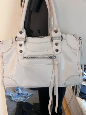 Vit Balenciagaliknande handväska - Säljer en vit handväska i glansigt skinn med silverfärgade detaljer och dragkedja framtill. Väskan har dubbla handtag och dekorativa nitar samt långa läderband. Perfekt storlek för det viktigaste och ger en cool vibe till din outfit.