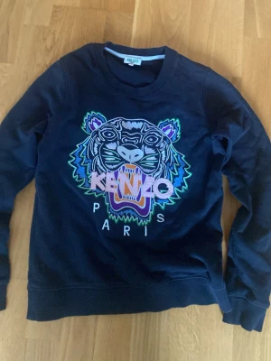 Svart sweatshirt med tiger från Kenzo - Snygg svart sweatshirt från Kenzo med det ikoniska broderade tigermotivet i flera färger och texten 'KENZO PARIS' på bröstet. Tröjan har rund halsringning och långa ärmar. Perfekt för dig som vill sticka ut med en statement-piece.