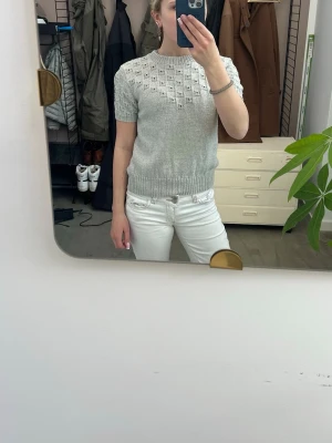 Ljusgrå stickad topp med kort ärm - Snygg ljusgrå stickad topp med korta ärmar och rund hals. Tröjan har ett diskret hålmönster upptill och bred ribbstickad mudd i midjan. Passar till både jeans och kjol.