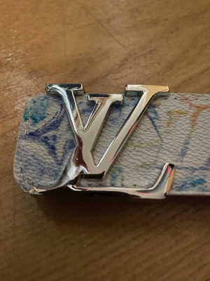  Vitt bälte med silver spänne - Snyggt vitt bälte med silverfärgat spänne. Bältet har färgglada detaljer i blått och orange på det präglade lädret. Perfekt accessoar för att lyfta din outfit.