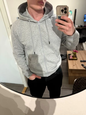 Ljusgrå zip-hoodie med huva - En stilren ljusgrå hoodie från Jack& Jones med dragkedja framtill, huva med dragsko och två stora fickor. Tillverkad i mjuk bomullsblandning och har ribbade muddar vid ärmslut och nederkant. Perfekt för en avslappnad och trendig look.