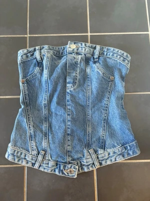Blå jeans tubtopp med dragkedja - Säljer en unik blå tubtopp i denim med klassiska jeansdetaljer som bälteshällor, knappar och fickor. Toppen har dragkedja och smock baktill för extra passform. Perfekt för dig som vill sticka ut med en edgy och trendig look.