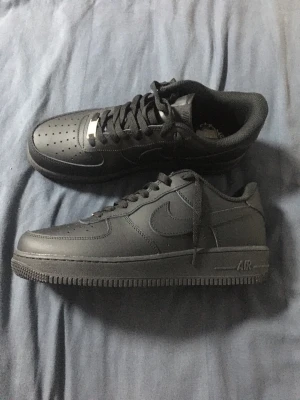 Nike Air Force 1 helsvarta sneakers - Säljer ett par klassiska Nike Air Force 1 i helsvart läder. Skorna har lågt skaft, perforerad tåbox och ikonisk swoosh på sidan. Tjock sula med AIR-detalj och svarta skosnören med metallbricka framtill. Perfekta för dig som gillar stilrena och tidlösa sneakers.