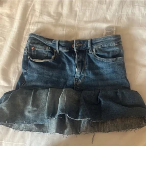 Jeanskjol med volangkant - Säljer en blå jeanskjol med volangkant och råa kanter. Kjolen har klassiska fickor fram och bak samt dragkedja och knapp framtill. Perfekt för dig som gillar en avslappnad och trendig stil.
