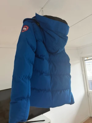 Blå vinterjacka från Canada Goose - Säljer en blå dunjacka från Canada Goose med huva och klassisk patch på ärmen. Jackan är tjockt vadderad och har en sportig look, perfekt för kalla vinterdagar. Materialet är slitstarkt och jackan har en modern, puffig passform.