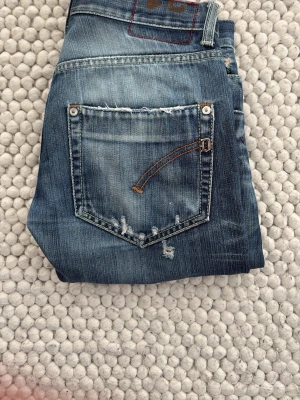 Dondup jeans  - Säljer ett par blå jeans från Dondup med slitningar och distressed detaljer på både ben och bakficka. Jeansen har raka ben och klassisk femficksdesign. Materialet är denim och färgen är en tvättad blå nyans med ljusare partier. 