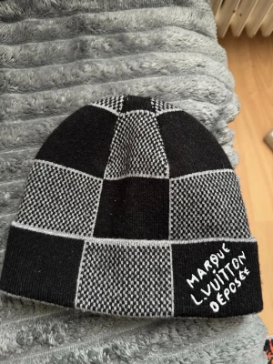 Svart och grå rutmönstrad mössa  - Cool mössa med rutmönster i svart och grått. Stickad design med broderad text på kanten. Perfekt för dig som vill ha en unik accessoar med streetst.