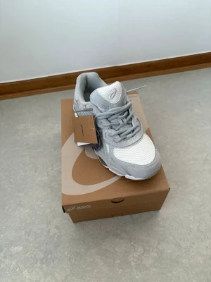 Gråvita ASICS sneakers mesh & skinn - Snygga sneakers från ASICS i grått och vitt med coola detaljer. Ovandelen är i mesh och syntetiskt skinn för en sportig vibe. Skorna har en chunky sula och klassisk snörning. Perfekta för dig som gillar streetstyle och bekväma skor.