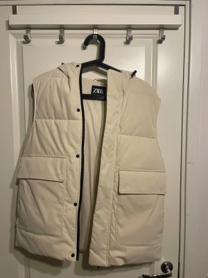 Beige dunväst med huva från Zara - Stilren beige dunväst från Zara med huva och stora fickor framtill. Västen har quiltad design, tryckknappar och är tillverkad i ett mjukt, vadderat material som håller värmen. Perfekt för lager-på-lager och enkel att matcha med olika outfits.