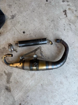 Avgassystem till moped, kolfiber och metall - Säljer ett avgassystem till moped med ljuddämpare i kolfiber och böjd avgasrör i metall. Systemet har svetsade sektioner och medföljer fäste och klämma. Passar dig som vill uppgradera ljud och prestanda på din moppe.
