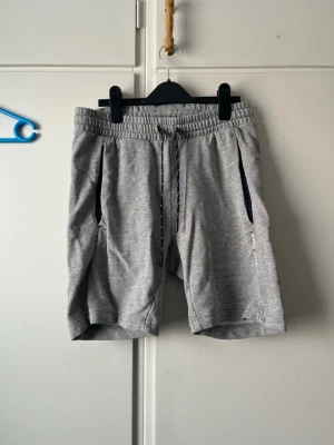 Grå mjukisshorts med snörning - Snygga grå mjukisshorts med elastisk midja och snörning framtill. Shortsen har två fickor med mörkblå detaljer och är perfekta för en avslappnad stil. Tillverkade i mjukt material som känns skönt mot huden.