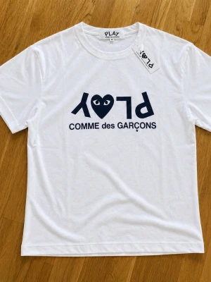  Cdg t-shirt - Snygg vit t-shirt från Comme des Garçons PLAY med det ikoniska hjärtloggan och texten 'PLAY' tryckt i svart på bröstet. Klassisk passform och rund hals. Perfekt för dig som gillar streetwear och vill ha något stilrent men ändå unikt.