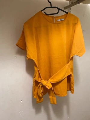 Gul topp med knyt från Pieces - Snygg gul topp från Pieces i storlek XS. Modellen har korta ärmar, rund halsringning och en trendig knytning i midjan. Perfekt för att liva upp din outfit med en poppig färg och cool detalj. Materialet känns lätt och luftigt.