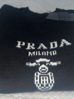 Svart stickad tröja från Prada - Svart stickad tröja från Prada med vit logotyp och Milano-tryck framtill. Tröjan har en klassisk rund halsringning och ett coolt grafiskt mönster under loggan. Perfekt för dig som gillar exklusiva streetwear-vibbar.
