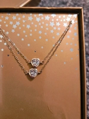 Guldigt halsband med hjärtan x2 - Säljer ett set med 2 likadana guldiga halsband med varsitt hjärtformat hänge med glittrande stenar. Kedjan är tunn och elegant, och halsbandet kommer i en snygg guldfärgad presentask med stjärnmönster. Perfekt accessoar för att lyfta vilken outfit som helst. Perfekt som vänskap, systrar eller mor och dotter  Kan även hämtas I Partille  J