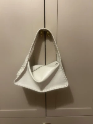 vit axelremsväska med flätad kant - snygg vit handväska i mjukt material med unik triangulär form. Väskan har en bred axelrem och dekorativ flätad söm längs kanterna. Stängs med dragkedja upptill och har en modern, stilren look. 