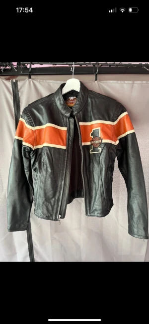 Harley-Davidson svart skinnjacka - Svart skinnjacka från Harley-Davidson med orange och beige ränder över bröst och rygg. Snyggt broderat Harley-Davidson-märke framtill och stort tryck på ryggen. Jackan har dragkedja framtill och långa ärmar. Perfekt för dig som gillar bikerstil. Mindre fläck på axel, se bild
