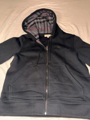 Svart Burberry hoodie med dragkedja - Snygg svart hoodie från Burberry med dragkedja och känguruficka. Insidan av huvan har klassiskt rutigt Burberry-mönster i grått, svart och rött. Tillverkad i mjuk bomull, perfekt för en chill och stilren look.
