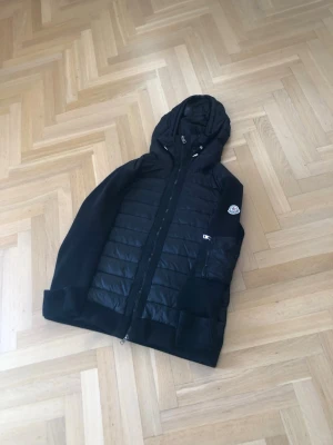 Moncler cardigan - Snygg svart cardigan från Moncler med quiltad front, stickade ärmar och huva. Jackan har dragkedja framtill, ribbade muddar och Moncler-logga på ärmen. 