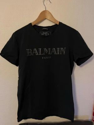 Svart Balmain Paris t-shirt - Snygg svart t-shirt från Balmain Paris med klassisk logga tryckt på bröstet. T-shirten har rund halsringning och korta ärmar. Perfekt för dig som gillar exklusiva streetwear-plagg och vill ha något stilrent från ett välkänt märke.