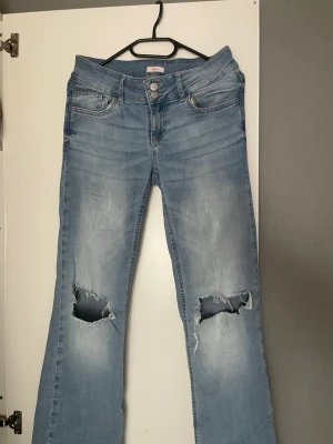 SLjusblå bootcut jeans med hål - Säljer ett par ljusblå bootcut jeans med slitna hål över knäna och klassiska fem fickor. Jeansen har dubbla knappar i midjan och bakfickor med lock och knapp. Perfekta för en avslappnad och trendig look. Lite slitna vid bensluten.